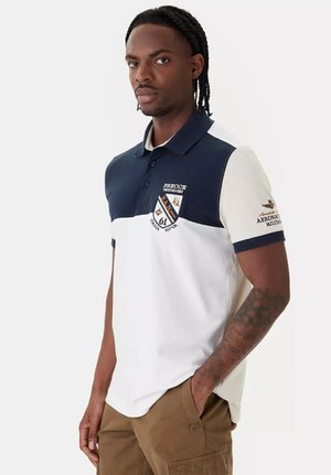 Uomo in piedi con una mano in tasca, indossa una polo bicolore con stemmi militari e testo sul petto e sulla manica.