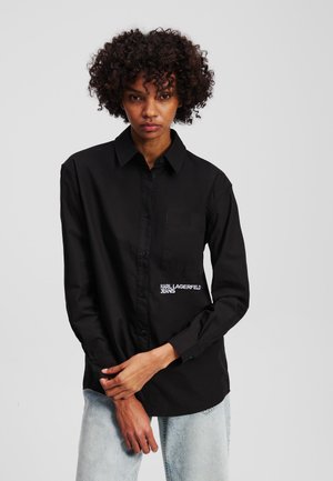 Zwarte long-sleeve shirt van glad materiaal met een knoopsluiting aan de voorkant, kraag, enkele borstzak en een wit geborduurd logo aan de zijkant.