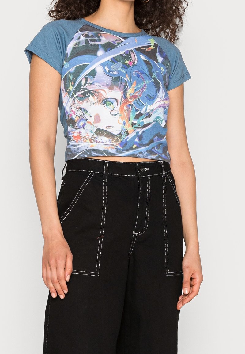 Blue raglan crop top featuring a colorful graphic print on the front. Paired with high-waisted black pants with white stitching details.
