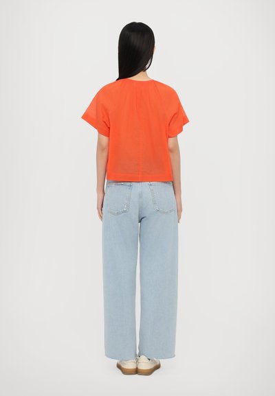Oranges T-Shirt mit kurzen Ärmeln und lockerer Passform, mit feiner Rückennaht; kombiniert mit hellblauen weit geschnittenen Jeans und beigen Schuhen.