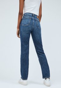 Jeans de denim azul con un diseño de pierna recta, que presentan una cintura alta, bolsillos traseros y una sutil textura deslavada. Combinados con zapatillas blancas.