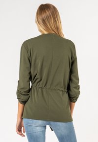 Olivgrön, långärmad topp med en avslappnad passform och rynkad midja. Tyget är mjukt med en slät yta. Bakvy från axel till fåll.