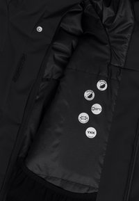 Svart jacka inuti som visar ett slätt, matt tyg med fem cirkelformade märken som anger vattentålighet, andningsförmåga, stretchfoder och YKK-dragkedjor.