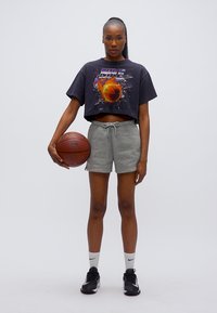 Μαύρο crop top T-shirt με ζωντανό γραφικό, γκρι σορτς, λευκές κάλτσες και μαύρα αθλητικά παπούτσια. Κρατώντας μια μπάλα μπάσκετ, στέκεται σε απλό φόντο.