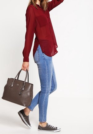 Maroon sheer button-up shirt, light blue skinny jeans, gray handbag, and gray sneakers. Hand raised, posing to the side.