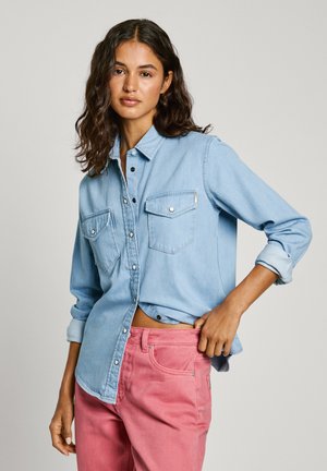Himmelblaues Jeanshemd mit zwei Brusttaschen, Knopfverschluss und hochgekrempelten Ärmeln, kombiniert mit rosa Hosen. Glatte Textur, lässiges Design.