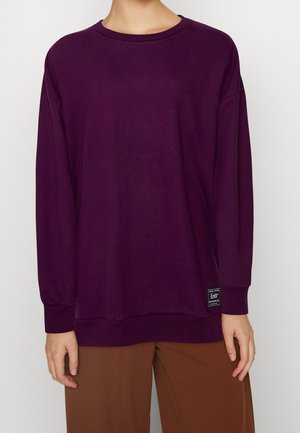 Person iført en ensfarvet, mørk lilla langærmet skjorte med brune bukser, stående mod en neutral baggrund.