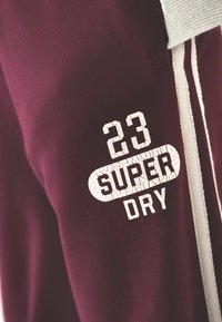 Felpa maroon con cappuccio e cordini bianchi che presenta del testo bianco screpolato "23 SUPER DRY" nella zona del petto.
