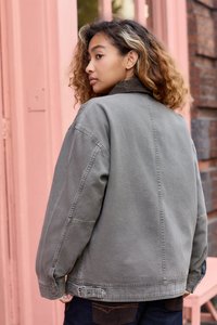 Gray denim jacket with a relaxed fit, featuring a pointed collar, side pockets, and adjustable strap at the hem. Textured fabric with visible seams.