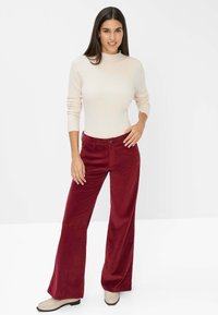 Rote Corduroy-Hose im weit geschnittenen Design mit vorderen Taschen, kombiniert mit einem hellbeigen, gerippten Pullover. Das Model trägt hellfarbige Loafers.