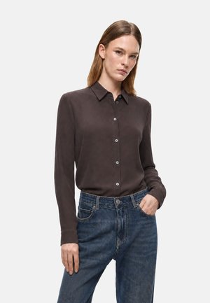 Donna che indossa una camicia marrone scuro con bottoni e maniche lunghe infilata in jeans blu a vita alta, con una mano in tasca.