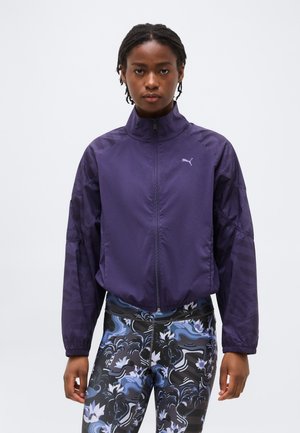 Naine, kandes lillat Puma lukuga jakki ja musta, sinise ja valge mustriga retuuse, seisab tavalise tausta ees.