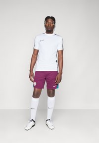 Jersey Nike a maniche corte bianco con accenti neri, pantaloni corti bordeaux con bande turchesi, calze bianche e scarpe da calcio in bianco e nero.