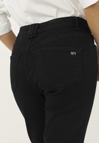 Persoană purtând blugi negri cu "IVY" brodat pe buzunarul din spate și o cămașă albă băgată în pantaloni.