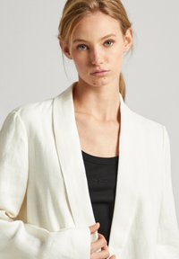 Blazer bianco in lino con vestibilità ampia, ampi rever e maniche lunghe, indossato su una canotta nera a coste con dettaglio del logo.