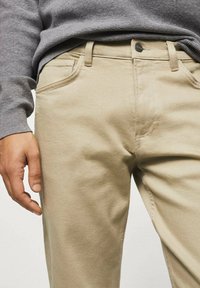 Chinos de algodón beige con corte recto, pasadores para cinturón y bolsillos laterales. Llevados con un suéter de punto gris que muestra una tela texturada.