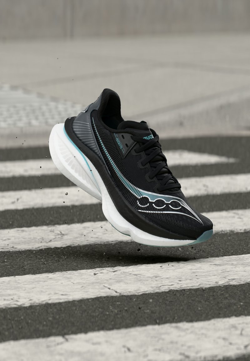 Μαύρο και τιρκουάζ αθλητικό παπούτσι με λευκή σόλα στον αέρα πάνω από διάβαση πεζών σε άσφαλτο, με ορατά σωματίδια βρωμιάς γύρω από τη σόλα.