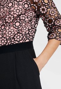 Robe noire avec une superposition en dentelle florale rose, présentant des manches trois-quarts et une taille ajustée, avec une texture lisse et des bords festonnés.