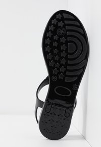 Semelle noire de sandale avec des motifs en forme d'étoile et courbes, debout sur un fond blanc.