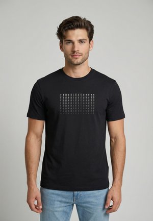 Junger Mann mit kurzem braunem Haar trägt ein schwarzes T-Shirt mit wiederholtem weißen "ARMANI EXCHANGE" Schriftzug und hellblaue Jeans, steht vor neutralem Hintergrund.