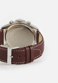 Reloj con correa de cuero marrón que presenta una caja de acero inoxidable, acentos plateados y un fondo grabado con el logo "TOMMY HILFIGER".