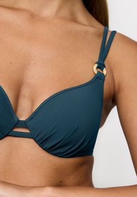 Bikini w kolorze turkusowym z gładkiej, elastycznej tkaniny, z dwiema cienkimi ramiączkami i złotym pierścieniem akcentującym na ramieniu.