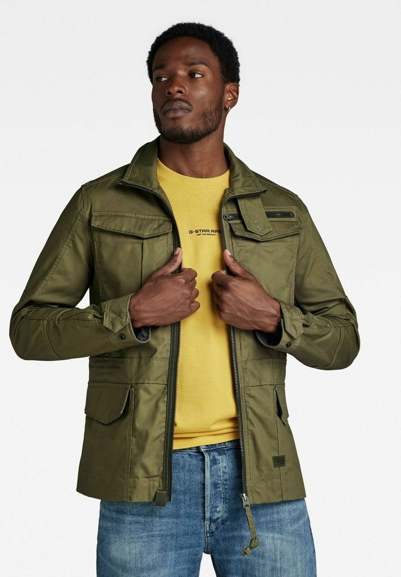 Giacca leggera verde oliva con zip e chiusura con bottone, dotata di diverse tasche frontali e un design casual, indossata sopra una camicia gialla.