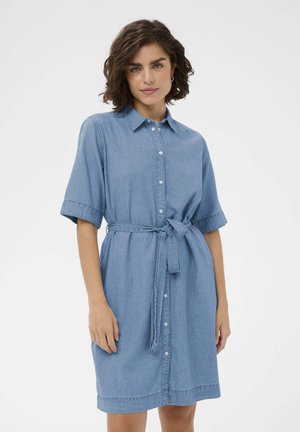 Kvinde stående iført en blå denimkjole med knapper, albuelange ærmer og et bindebånd i taljen mod en ensfarvet baggrund.