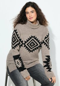 Beżowy sweter z golfem w czarne geometryczne wzory, w tym diamenty i zygzaki, ma długie rękawy i luźny krój.