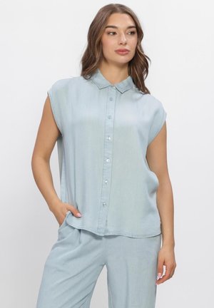Vrouw die een lichtblauw mouwloos blouse met knopen en bijpassende losse broek draagt, poseert met één hand in haar zak tegen een effen witte achtergrond.