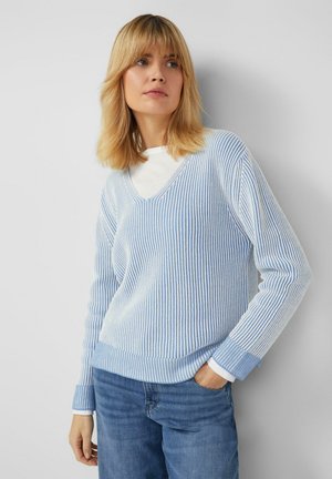 Femme blonde portant un pull à col en V rayé bleu clair et blanc sur une chemise blanche et un jean bleu, debout avec une main dans la poche.