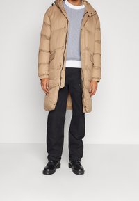 Beżowa kurtka puchowa z kapturem, posiadająca pikowany wzór, zamek błyskawiczny z przodu oraz zatrzaski. Nosi się ją na szarym swetrze i czarnych spodniach.