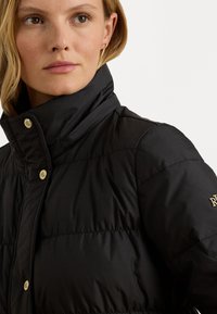 čierna pufová bunda s vysokým golierom, so zlatými cvokmi a logom na rukáve. Hladká textúra, ľahký dizajn.