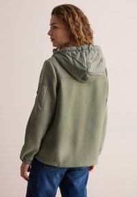 Giacca in pile verde oliva con cappuccio trapuntato a contrasto, polsini elasticizzati e tasca sul petto, caratterizzata da una vestibilità ampia e una texture sobria.