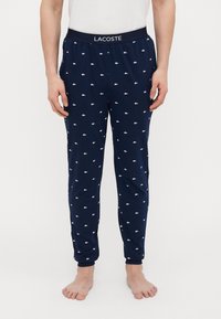 Marineblauwe pyjamabroek met witte Lacoste-logo's verspreid over de stof, elastische tailleband en taps toelopen ontwerp. Gemaakt van een zachte, lichte stof.