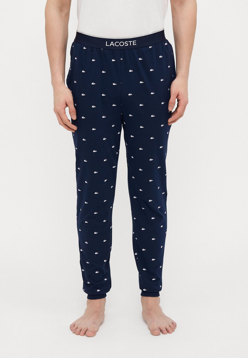 Marineblauwe pyjamabroek met witte Lacoste-logo's verspreid over de stof, elastische tailleband en taps toelopen ontwerp. Gemaakt van een zachte, lichte stof.