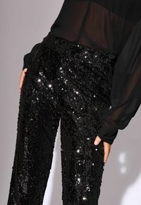Fekete, flitteres nadrág, texturált felülettel, testhezálló fazonnal és sima derékpánttal. Részben látható áttetsző fekete blúz.