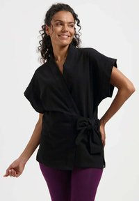 Schwarzes Wickeltop mit kurzen Ärmeln, einem gebundenen Taillendetail und lockerer Passform. Glatter Stoff ohne sichtbare Muster.