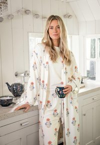 Frau in blumigen Schlafanzügen, die eine Tasse in einer hellen Küche hält und sich am Tresen neben einem blumigen Teekannen- und Teetassenset lehnt.