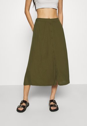 Femme portant une jupe midi boutonnée vert olive, un top court blanc et des sandales noires à boucle, debout les mains dans les poches de la jupe.