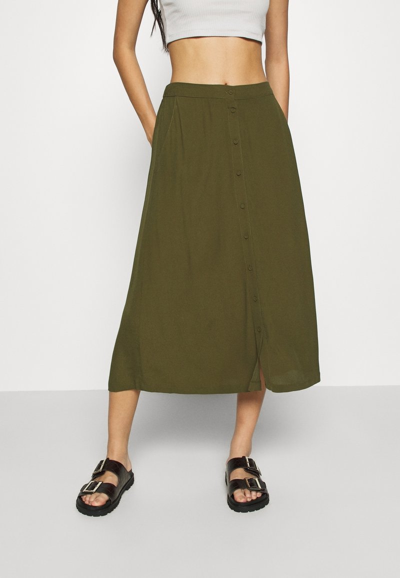 Femme portant une jupe midi boutonnée vert olive, un top court blanc et des sandales noires à boucle, debout les mains dans les poches de la jupe.