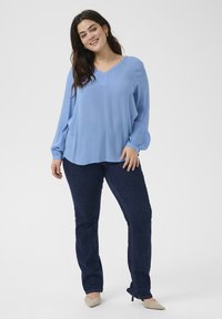 Blouse bleu clair à manches longues avec un col en V et un tissu subtilement texturé, associée à un jean droit bleu foncé et des chaussures beiges pointues.