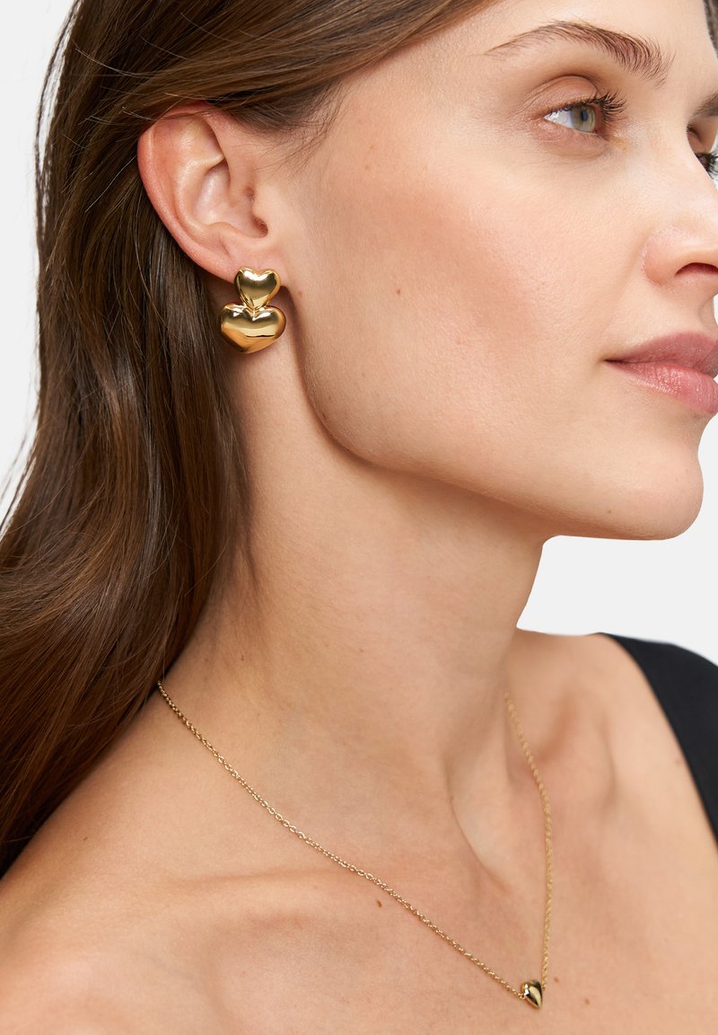 Goldene, herzförmige Ohrringe mit glänzender Oberfläche und eine zarte Kettenhalskette mit einem kleinen Heranhänger. Sanfte Texturen und elegantes Design.