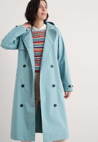 Jasnoniebieski płaszcz trench z podwójnym zapięciem, czarnymi guzikami i dużymi klapami, nałożony na sweter w wielokolorowe paski.