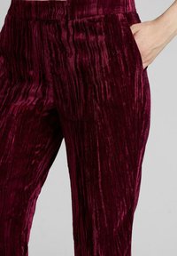 La mano derecha metida en el bolsillo de unos pantalones de terciopelo rojo oscuro brillante con textura, frente a un fondo simple.