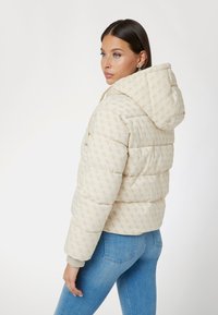 Eine Frau trägt eine cremefarbene, wattierte Kapuzenjacke mit dezentem Logo-Muster und blaue Jeans. Sie schaut über die Schulter vor einem einfarbigen Hintergrund.