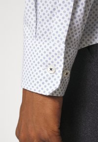 Manche de chemise blanche à poignets retroussés avec un petit motif floral bleu, deux boutons blancs, portée sur un poignet à peau foncée avec un pantalon en tissu foncé.