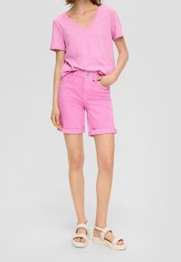 Mujer vistiendo una camiseta de V en rosa metida en unos pantalones cortos rosa con dobladillo enrollado, de pie con una mano en el bolsillo y sandalias trenzadas beige.