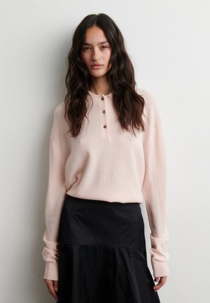 Vrouw met lang donker haar, gekleed in een lichtroze gebuttonneerde trui en een zwarte rok, staand tegen een effen achtergrond.