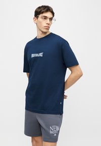 Νέος άνδρας με μπλε μπλουζάκι New Balance και γκρι σορτς στέκεται με τα χέρια πίσω από την πλάτη, κοιτώντας προς το πλάι σε απλό φόντο.